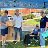 Four people standing outside under blue tent with stack of boxes and one man holding a box.