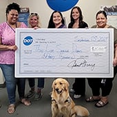 People in office building holding large check for $15,000 to Thin Line Service Dogs with golden retriever sitting under check.